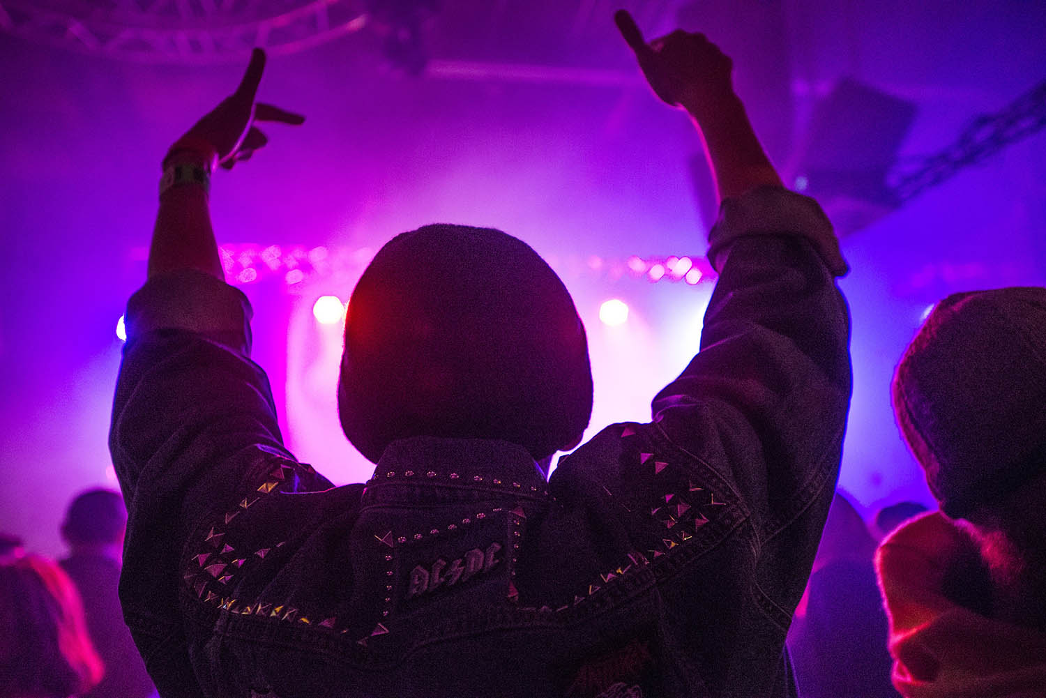 Back of a man at a concert. Hes wearing a beanie hat and a baggy denim jacket with studs. Photo with a purple hue